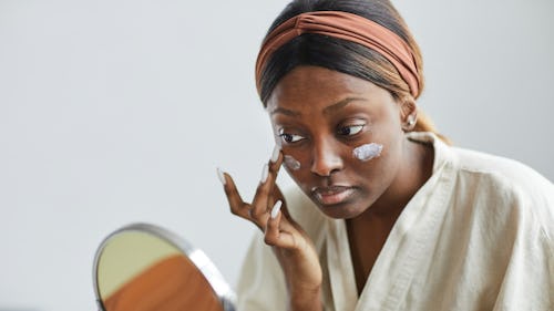 Image of a dark-skinned lady looking at a mirror while applying a skincare product on her face