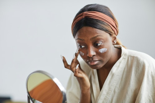 Image of a dark-skinned lady looking at a mirror while applying a skincare product on her face