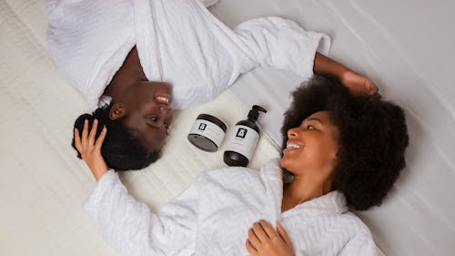 Image of two ladies lying on a bed with two skincare products placed on the bed
