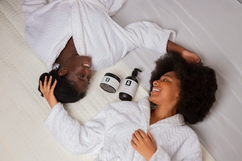 Image of two ladies lying on a bed with two skincare products placed on the bed