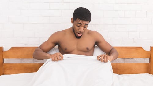 A topless black man sitting on a bed with a white duvet looking down to his penis