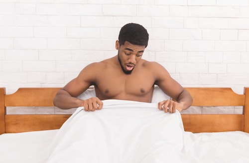 A topless black man sitting on a bed with a white duvet looking down to his penis