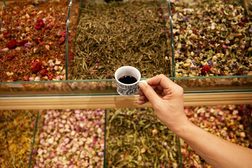 A picture showing a person carrying a cup of red zobo drink standing in front of a shelf containing herbs and leaves, including Hibiscus Sabdariffa