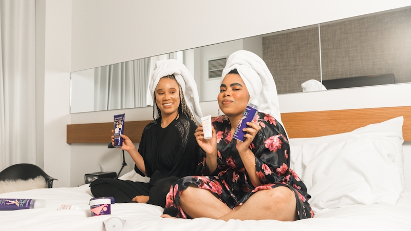 Two women sitting on a bed, holding and displaying different skincare products, including face creams
