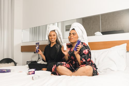 Two women sitting on a bed, holding and displaying different skincare products, including face creams