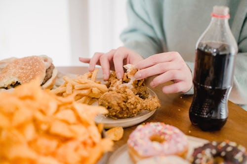 A girl eating large plate of fried chips, fried chicken, burgers, doughnuts and a bottle of soda