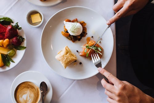 Image of a person eating small food portion with fork and knife on a table with a plate of fruits