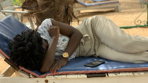Image of a lady with afro hair sleeping on a wooden long chair under a coconut tree