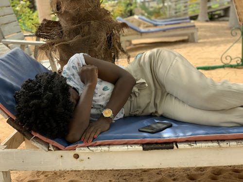 Image of a lady with afro hair sleeping on a wooden long chair under a coconut tree