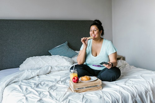 Image of an overweight woman on a bed eating food, and apple drinking from a glass of juice placed on the bed