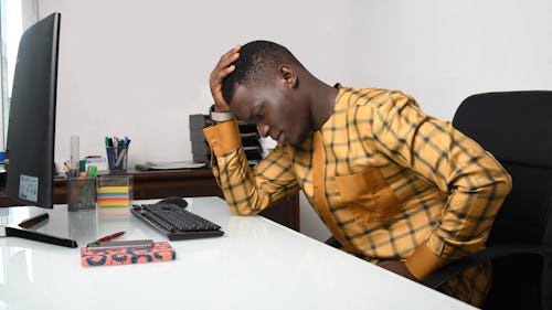 A Black man sitting on an office desk with hand placed on his head, looking tired and exhausted