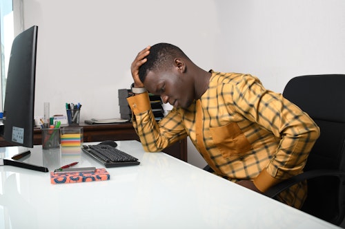 A Black man sitting on an office desk with hand placed on his head, looking tired and exhausted