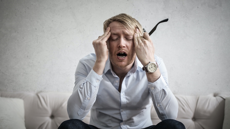 A stressed white man wearing a wrist watch rubbing his forehead with his hands 