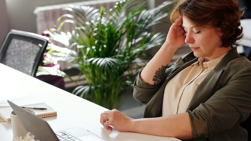 A woman feeling sick at work with hands placed on her forehead and a laptop placed on a table in front of her