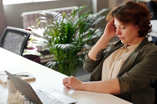 A woman feeling sick at work with hands placed on her forehead and a laptop placed on a table in front of her