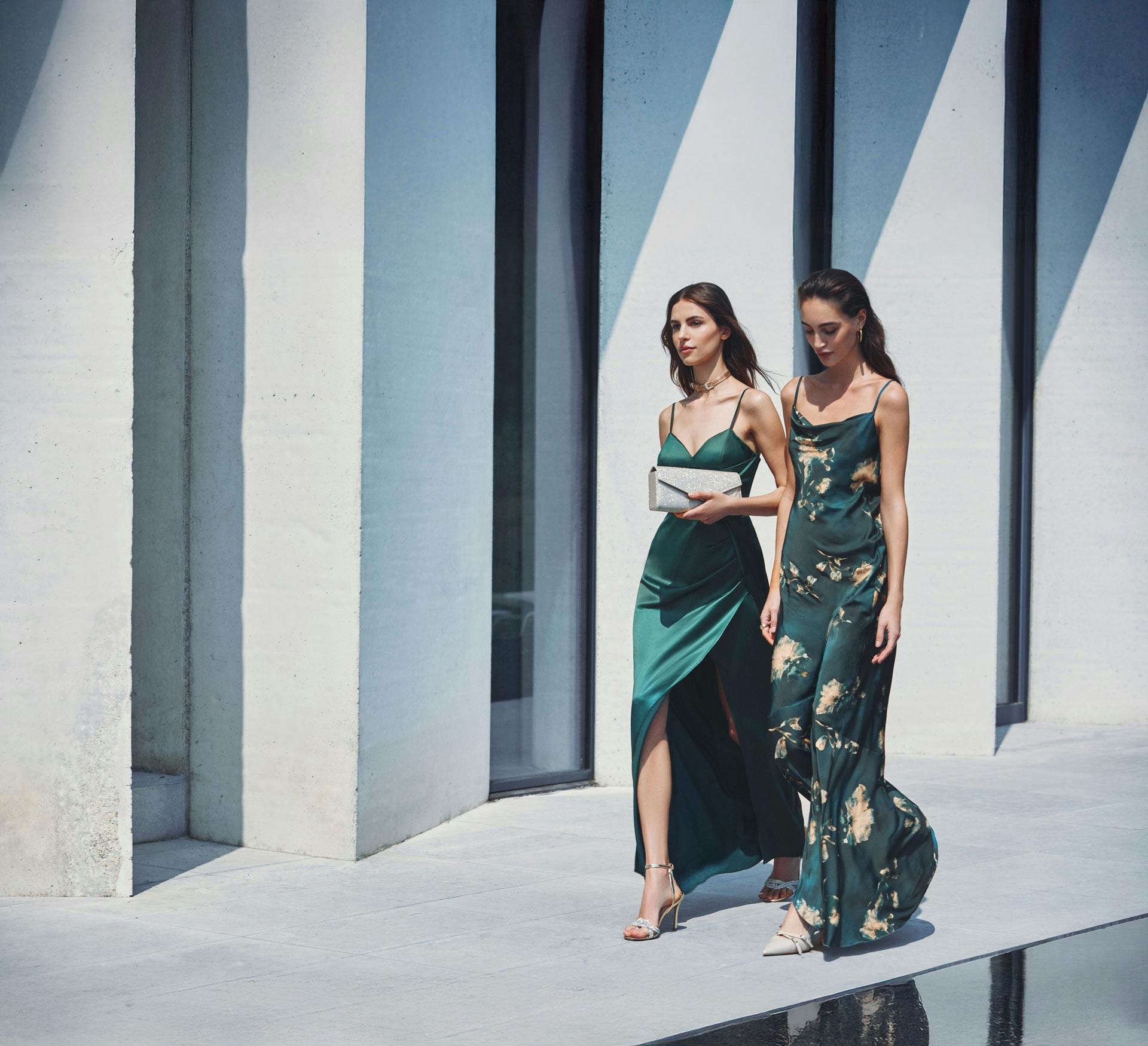 Due modelle indossano abiti da sera verde smeraldo, uno liscio e uno con stampa floreale, in location architettonica moderna con colonne bianche e blu