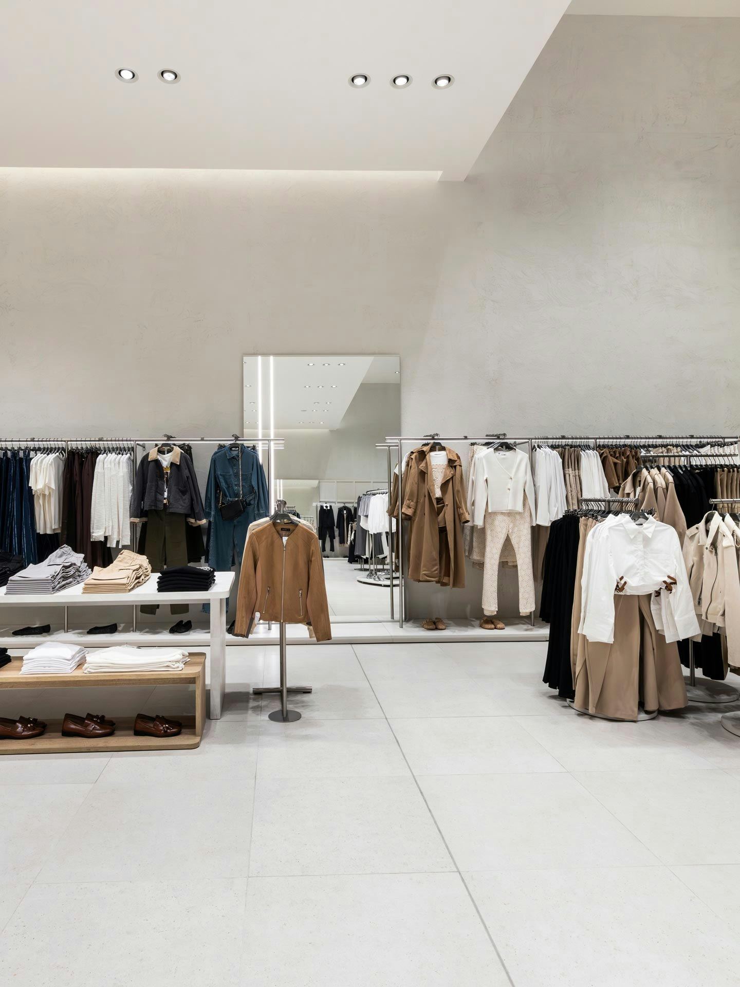 Interno moderno negozio Calliope con esposizione abbigliamento donna in tonalità neutre beige e marrone, tavoli centrali con capi piegati e appenderie perimetrali in ambiente minimalista bianco