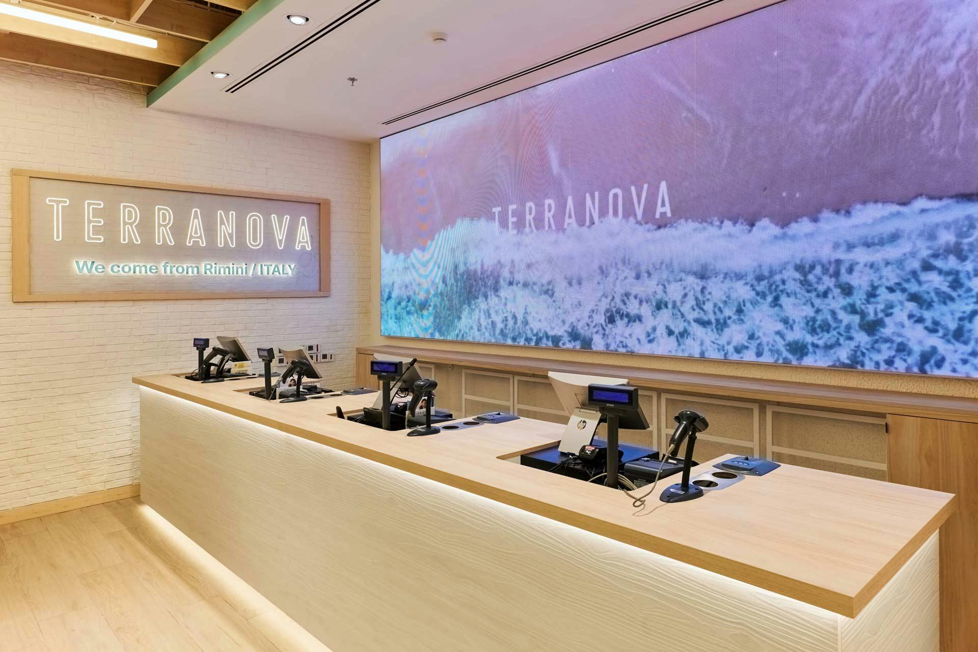 Area casse negozio Terranova con bancone in legno chiaro, LED wall che mostra immagini di onde marine e logo "We come from Rimini Italy"