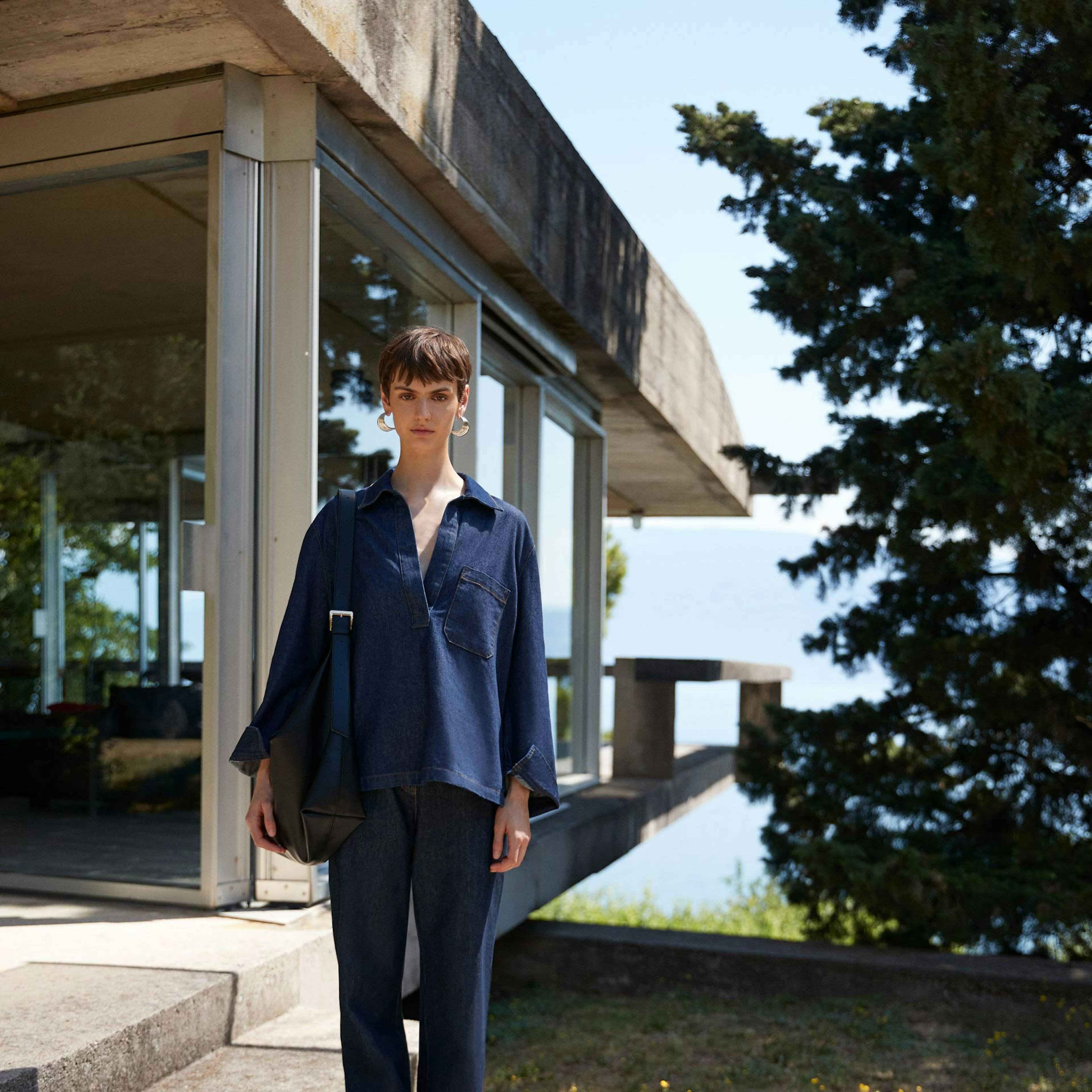 Modella indossa camicia blu oversize e pantaloni coordinati davanti a edificio in cemento brutalista, fotografia di moda outdoor con natura sullo sfondo