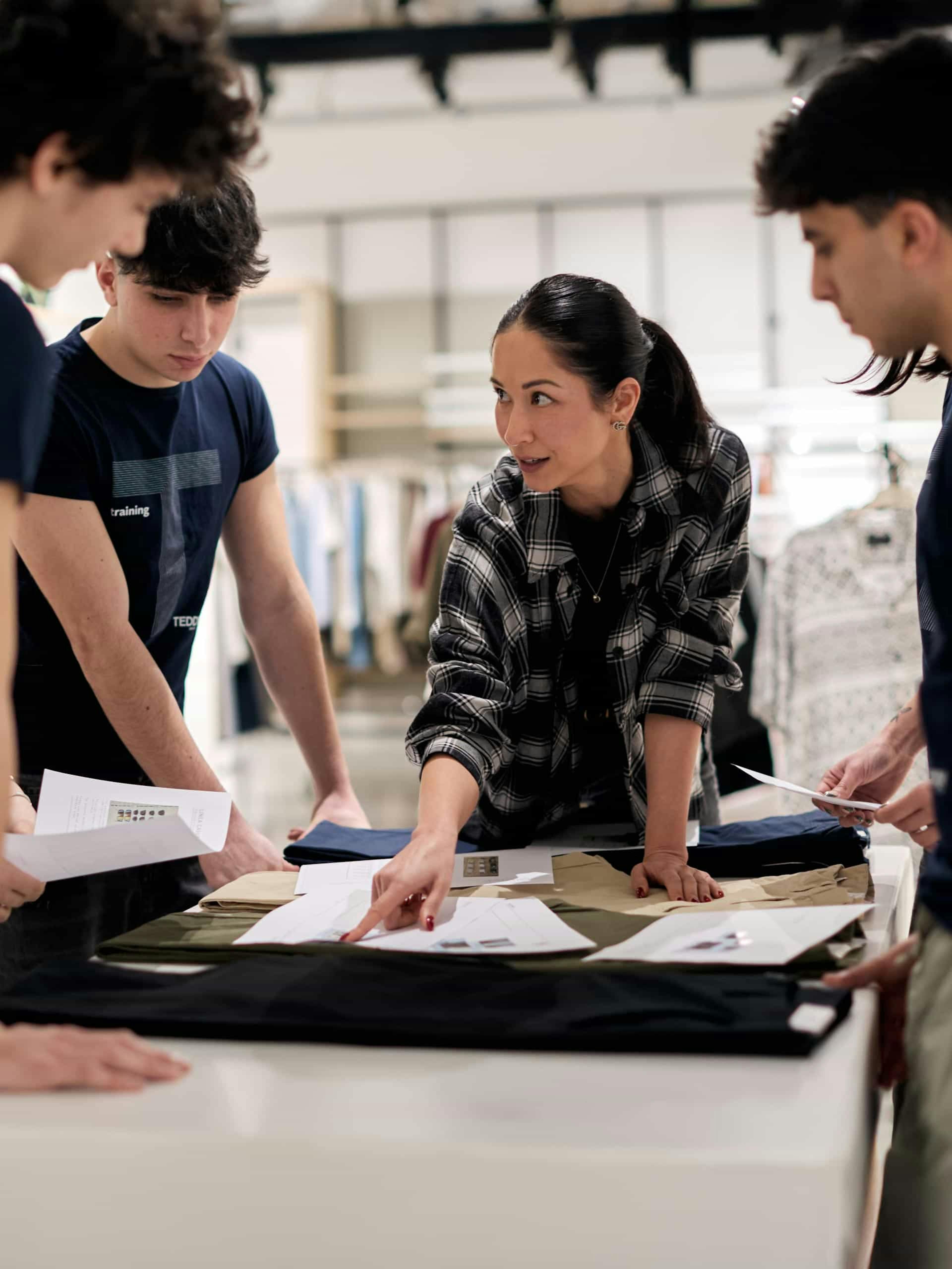 Team di design al lavoro su progetto moda con insegnante che guida studenti nella revisione di campioni tessuto e documenti su tavolo da lavoro