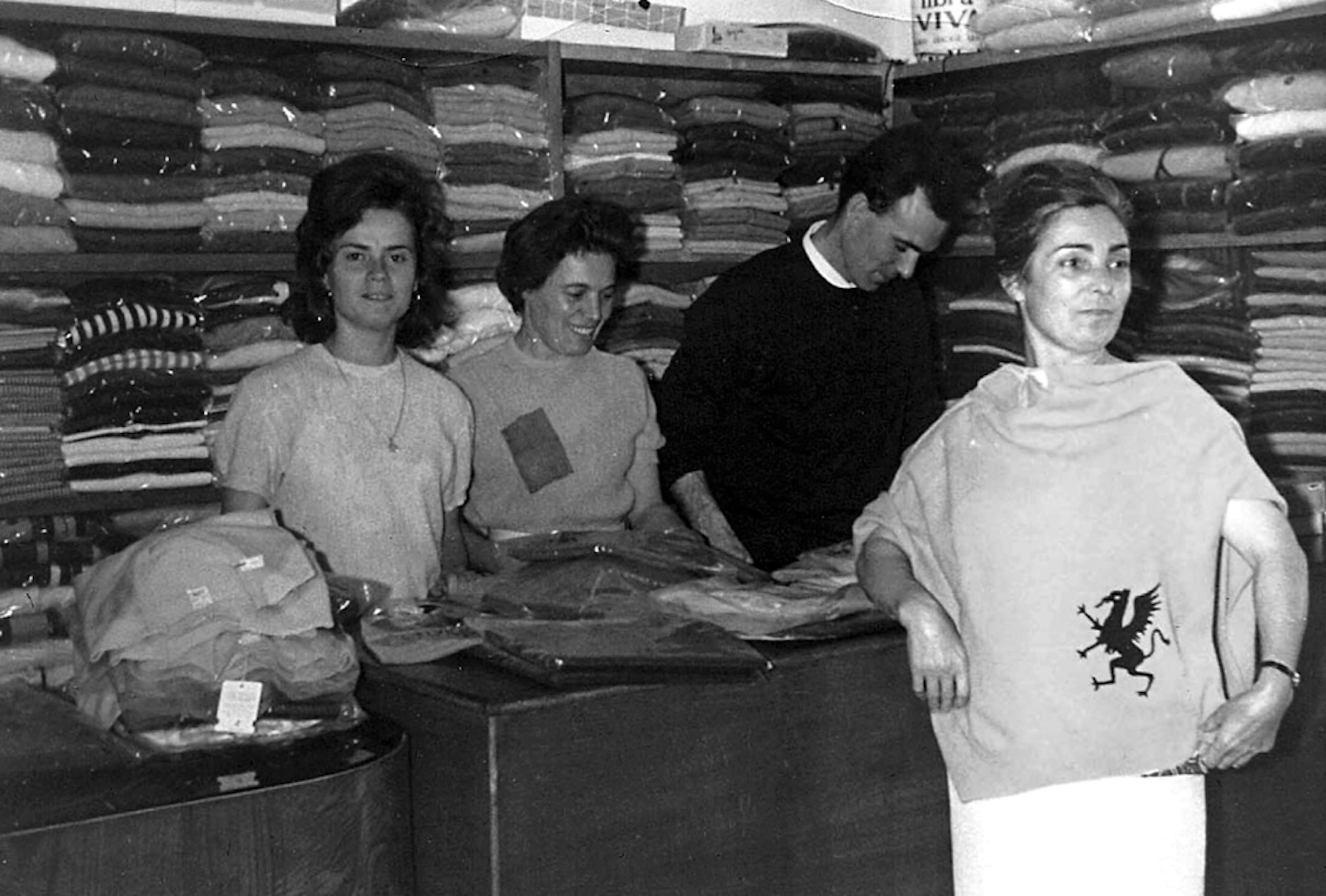 Foto storica in bianco e nero di un negozio di abbigliamento anni '60 con quattro persone dietro al bancone e scaffali pieni di maglioni sullo sfondo