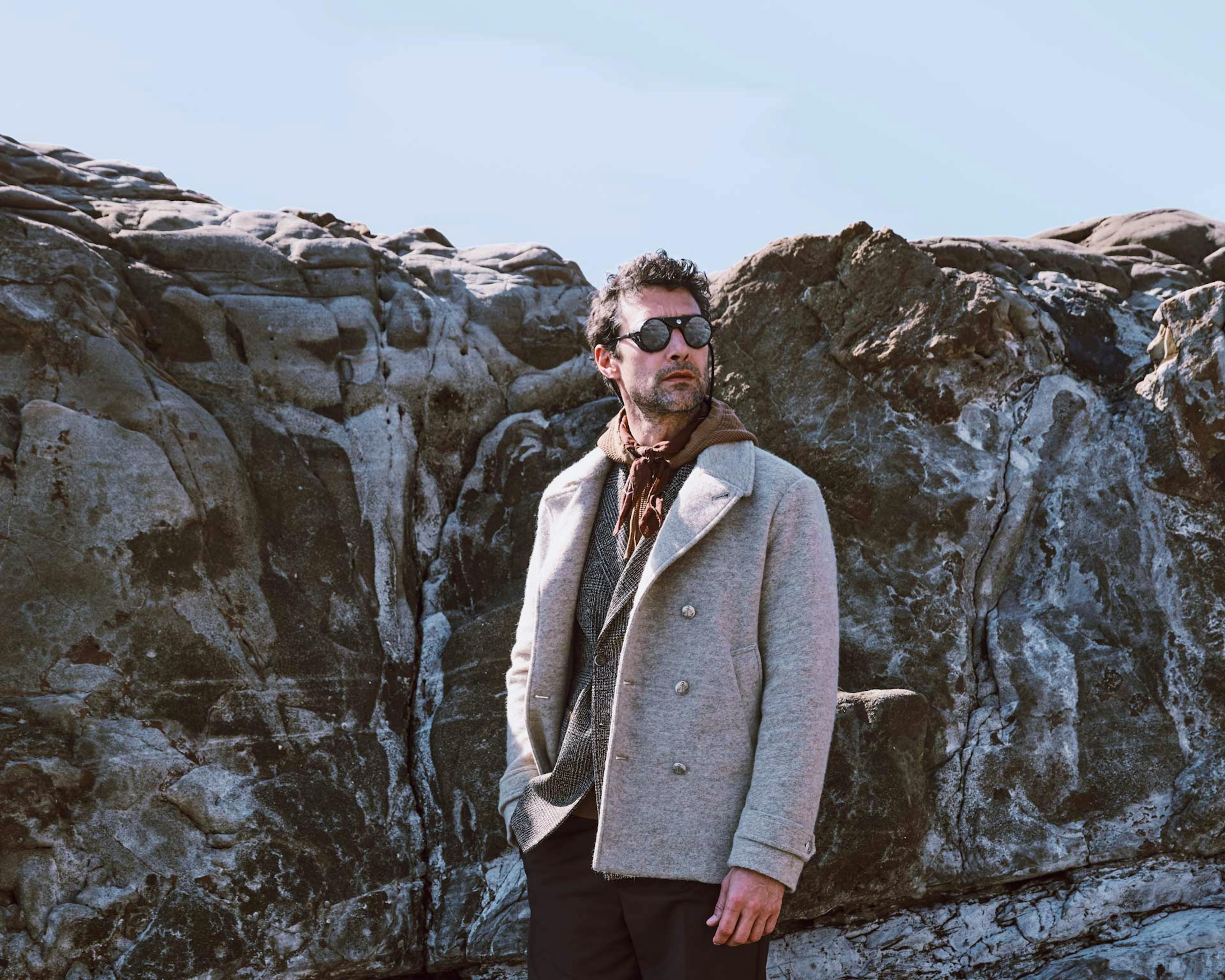 Modello maschile con occhiali da sole indossa cappotto doppiopetto grigio chiaro e sciarpa marrone, fotografato contro formazioni rocciose naturali sotto cielo azzurro
