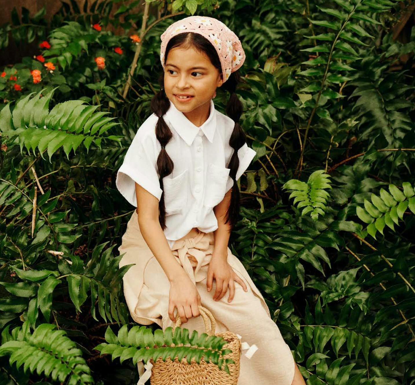 Bambina con bandana rosa e camicia bianca posa con borsa in paglia in giardino tropicale, immagine dal Report 2023 sulla sostenibilità