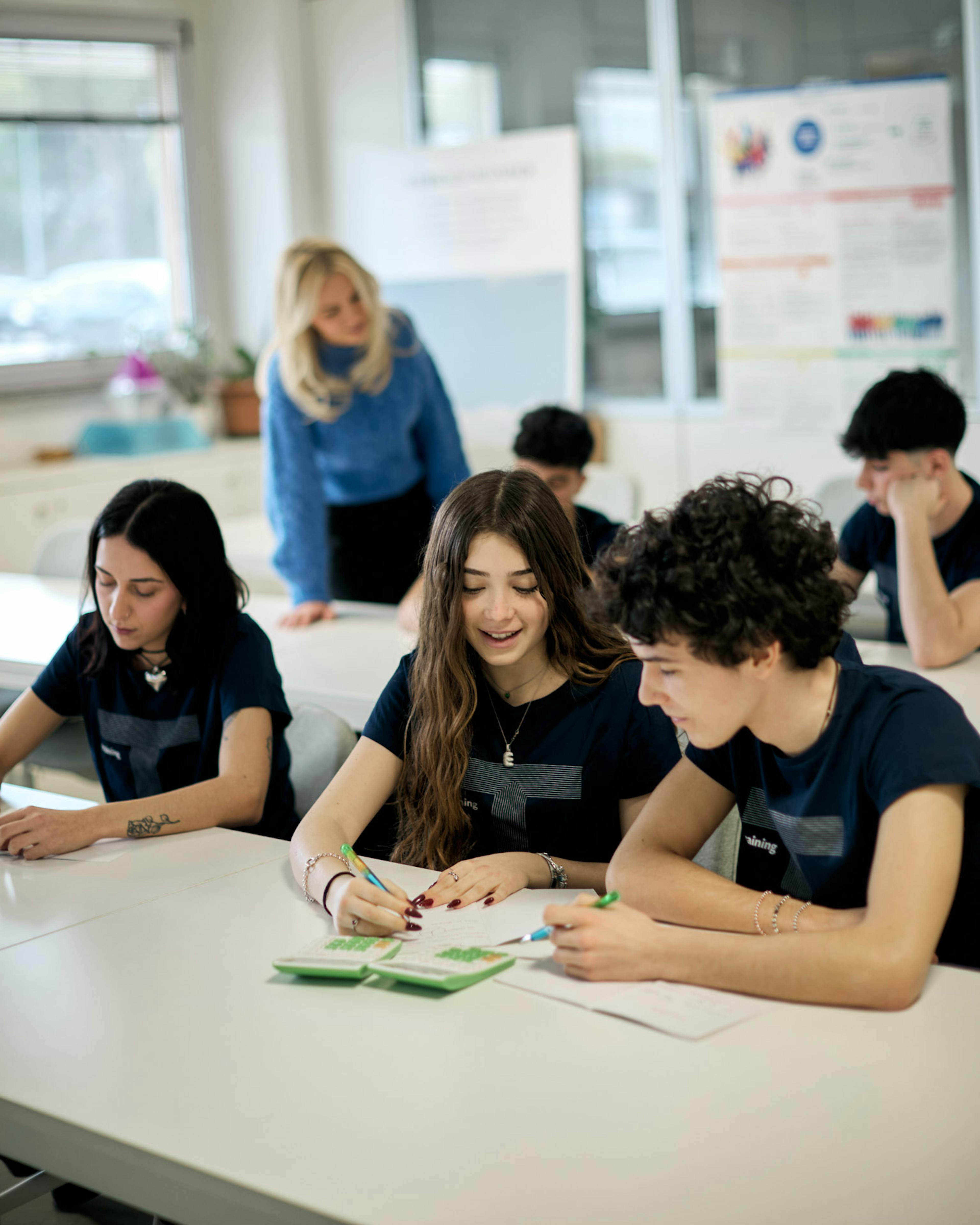 Studenti in aula didattica lavorano su progetti di fashion design, con insegnante bionda che supervisiona il lavoro di gruppo
