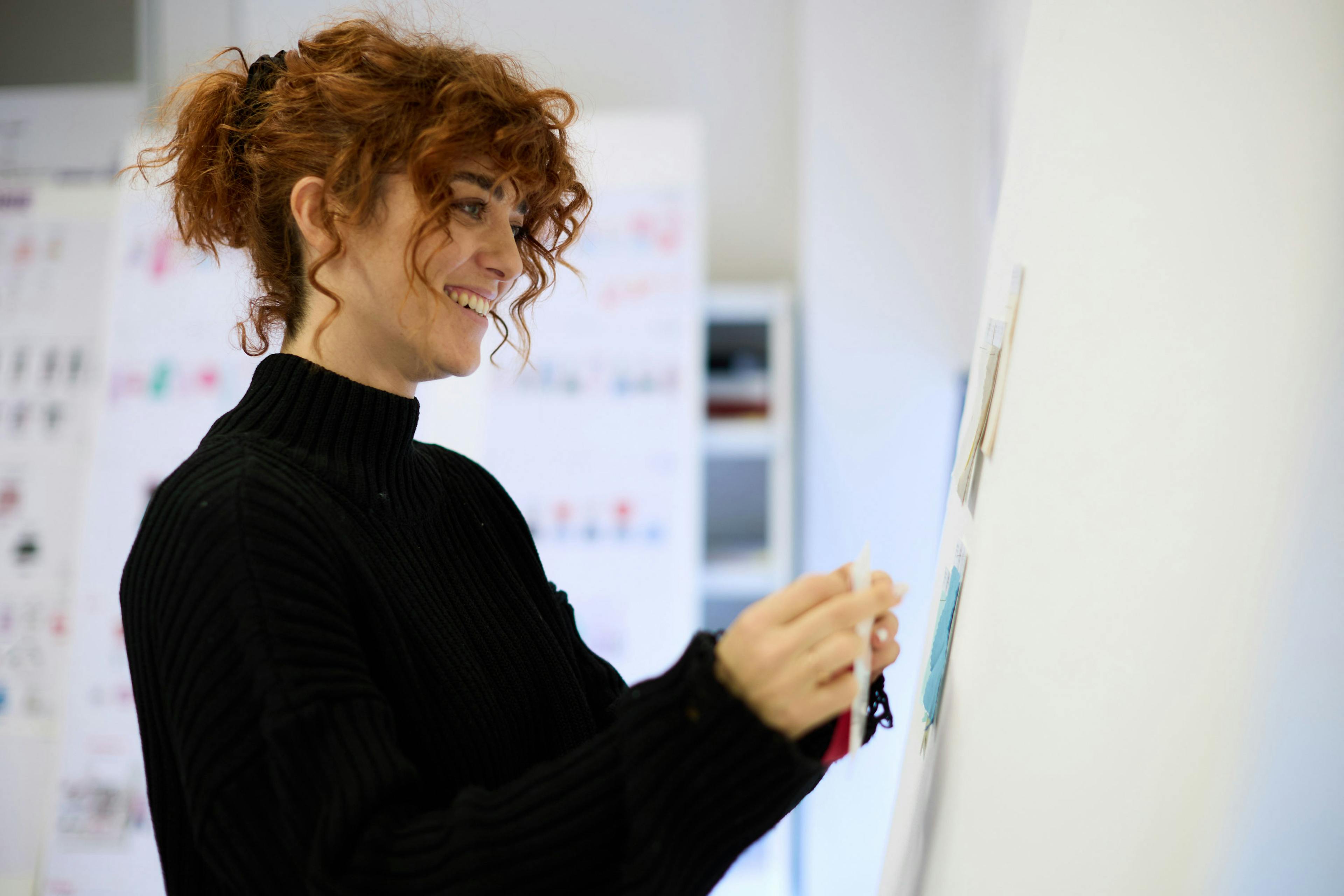 Designer con capelli ricci rossi sorride mentre lavora su lavagna con campioni colore in ufficio creativo di moda