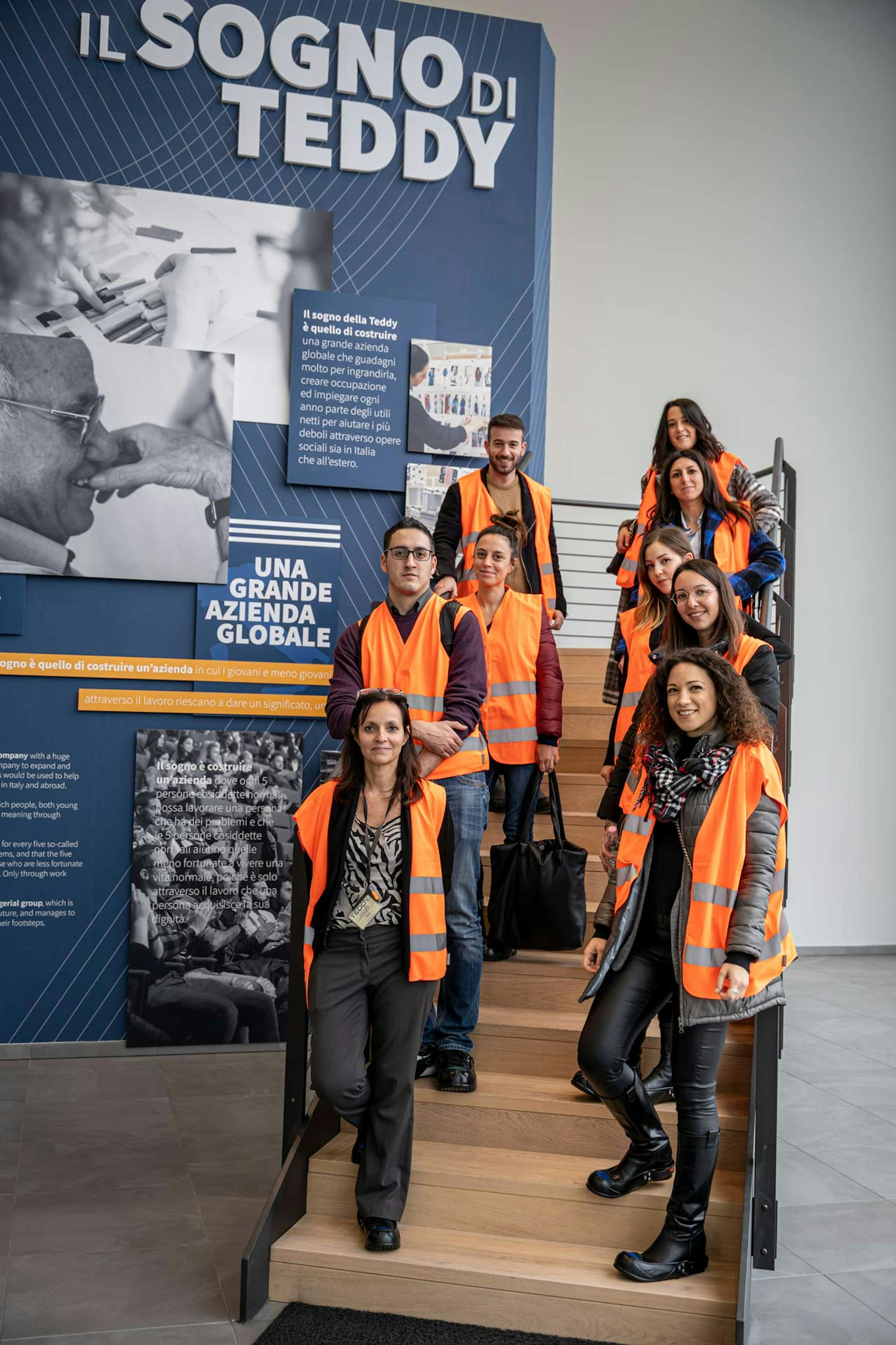 Gruppo di dipendenti Teddy con gilet ad alta visibilità arancioni su scala aziendale, pannello espositivo con mission "Il Sogno di Teddy"