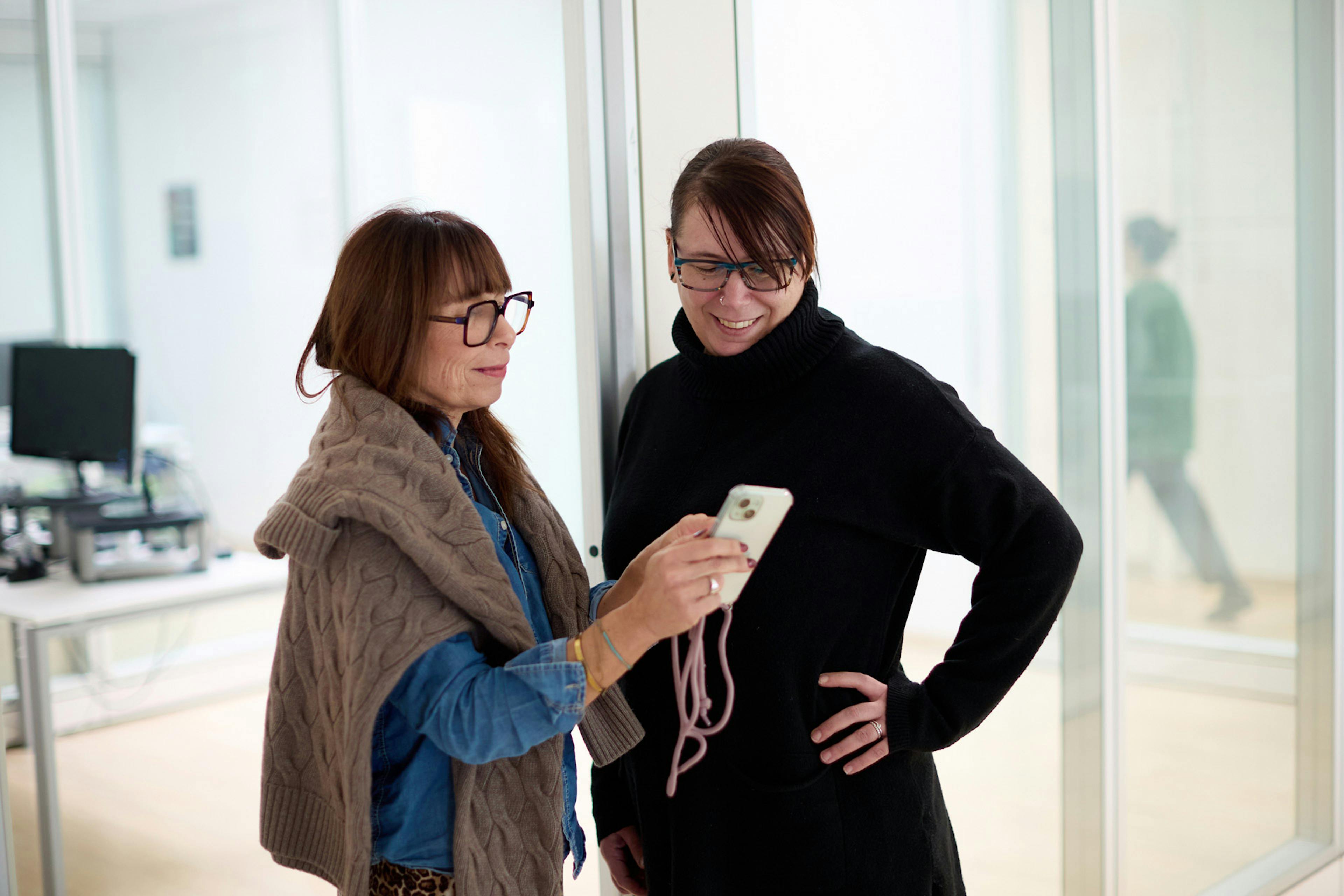 Due colleghe sorridenti in ufficio moderno condividono contenuti su smartphone, ambiente luminoso con grandi vetrate