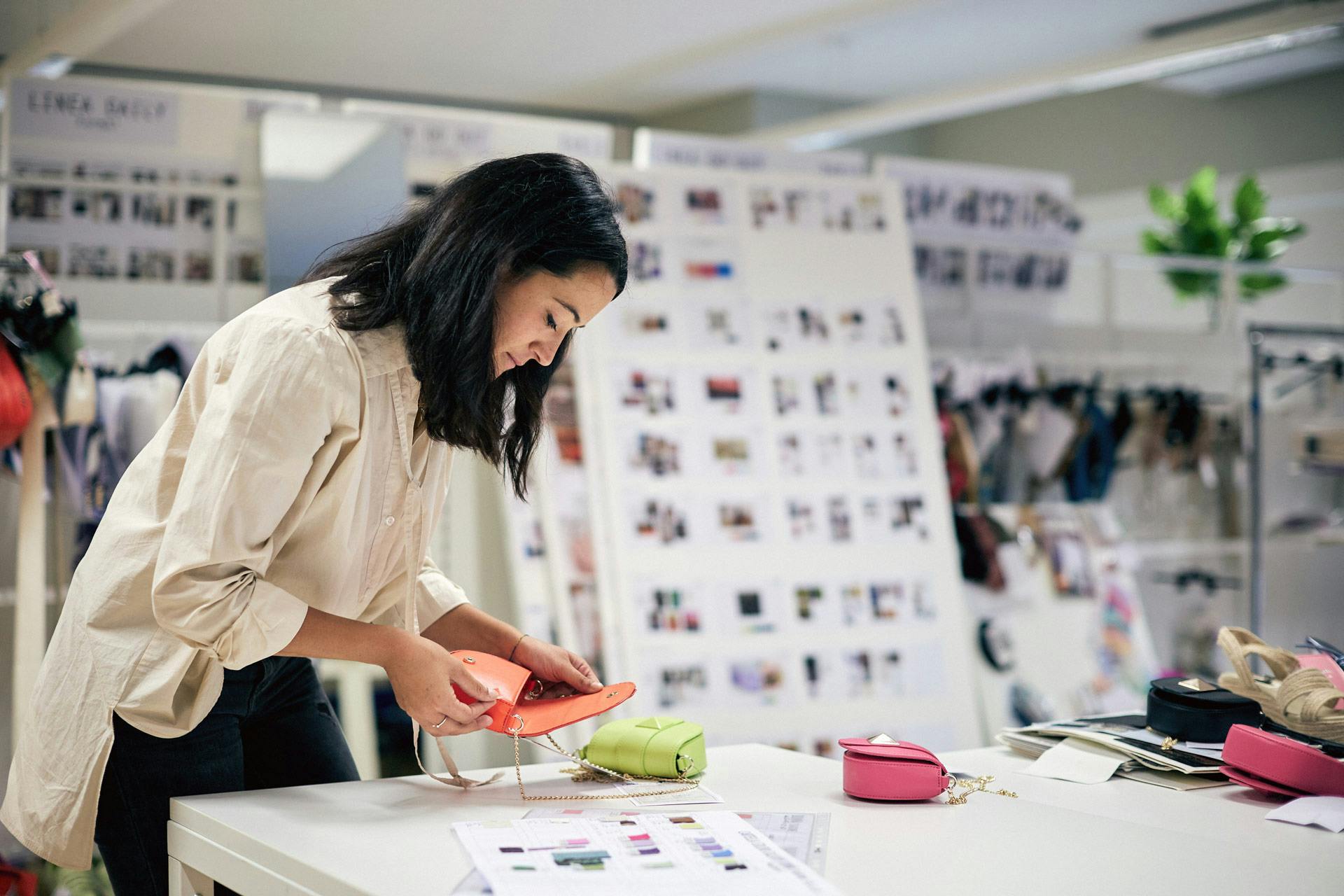 Designer donna esamina borse colorate rosa e verde su tavolo bianco con moodboard design e campioni colore alle spalle