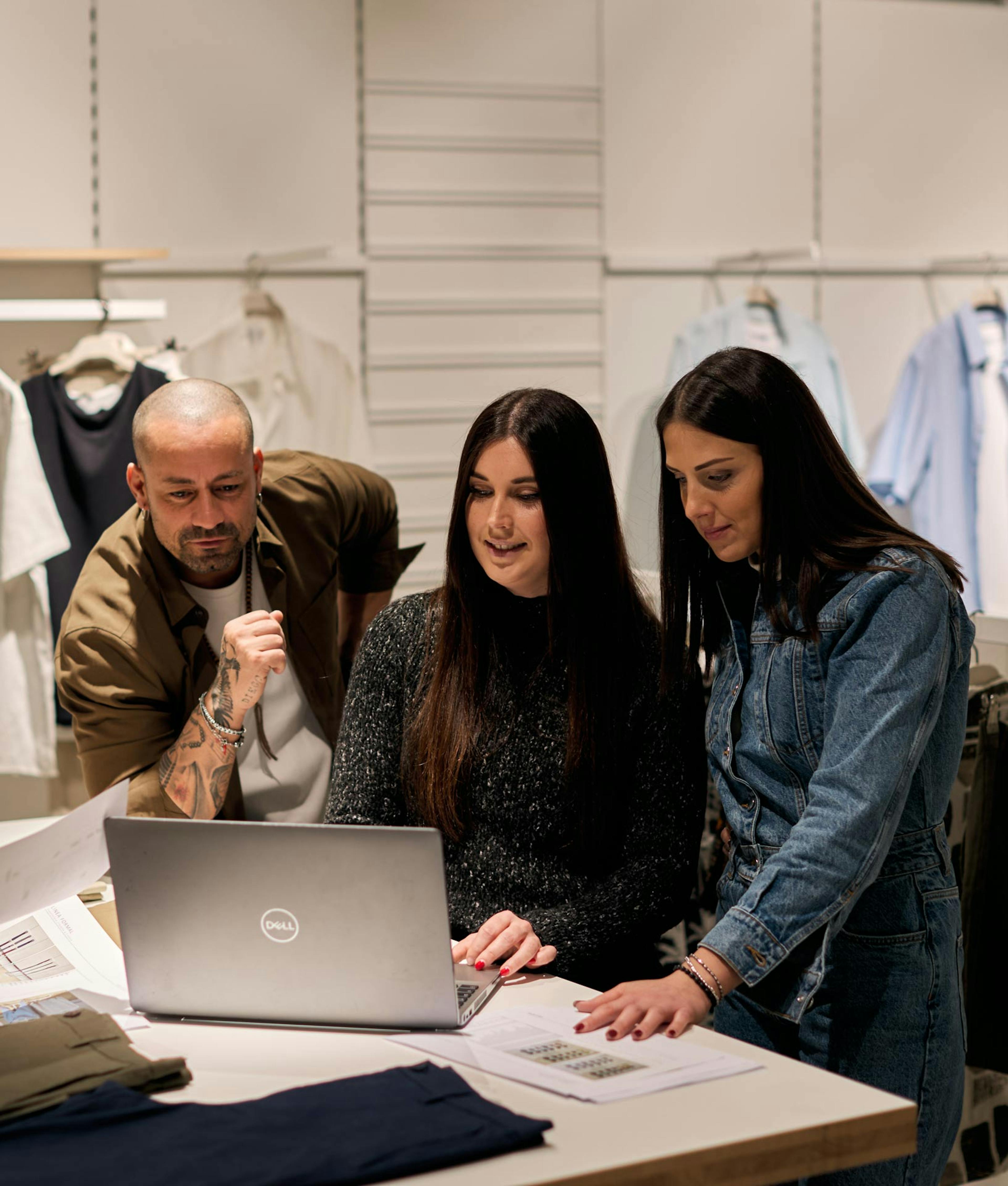Team di design moda collabora con laptop Dell in showroom, donna in maglione nero al centro tra due colleghi, abiti appesi sullo sfondo
