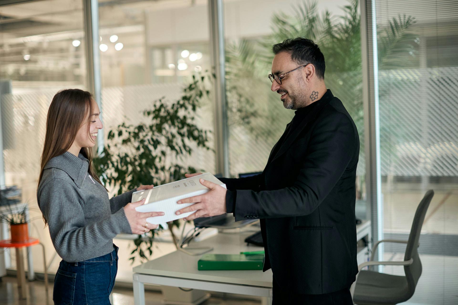 Due colleghi sorridenti si scambiano un pacco bianco in ufficio moderno con piante verdi e scrivania grigia