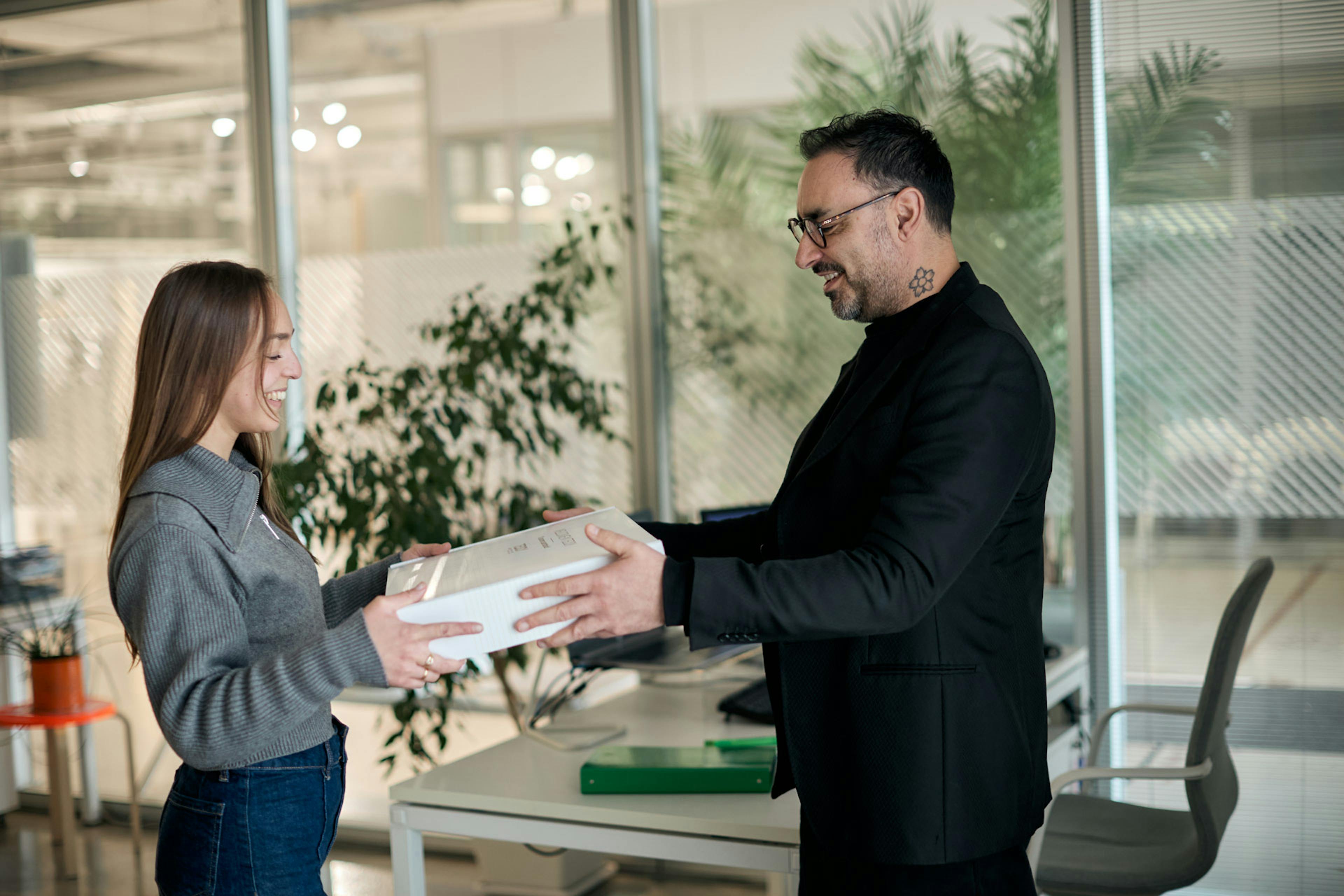 Due colleghi sorridenti si scambiano un pacco bianco in ufficio moderno con piante verdi e scrivania grigia