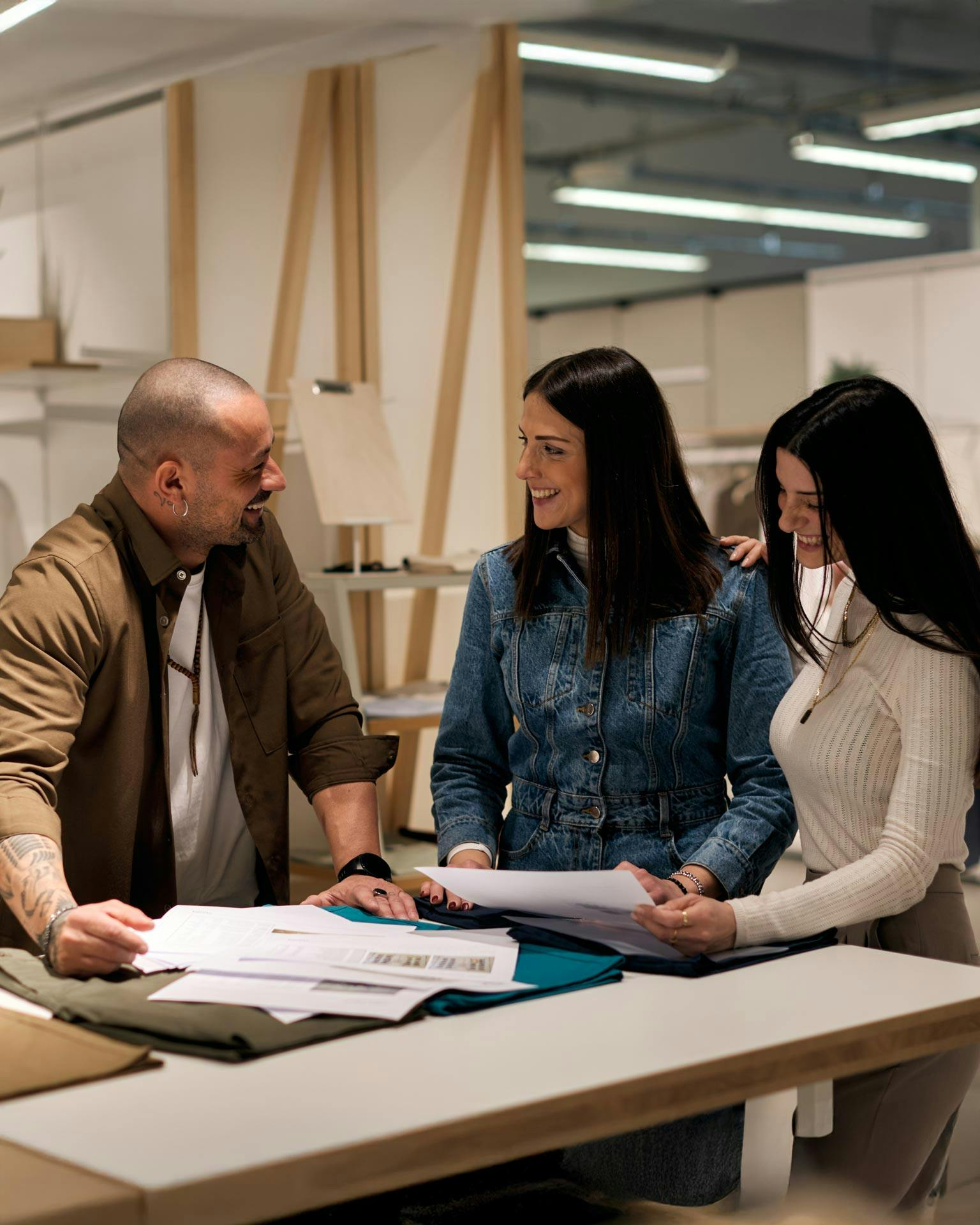 Tre professionisti della moda in studio esaminano campioni di tessuto su tavolo bianco, uomo con bomber beige e due donne in denim