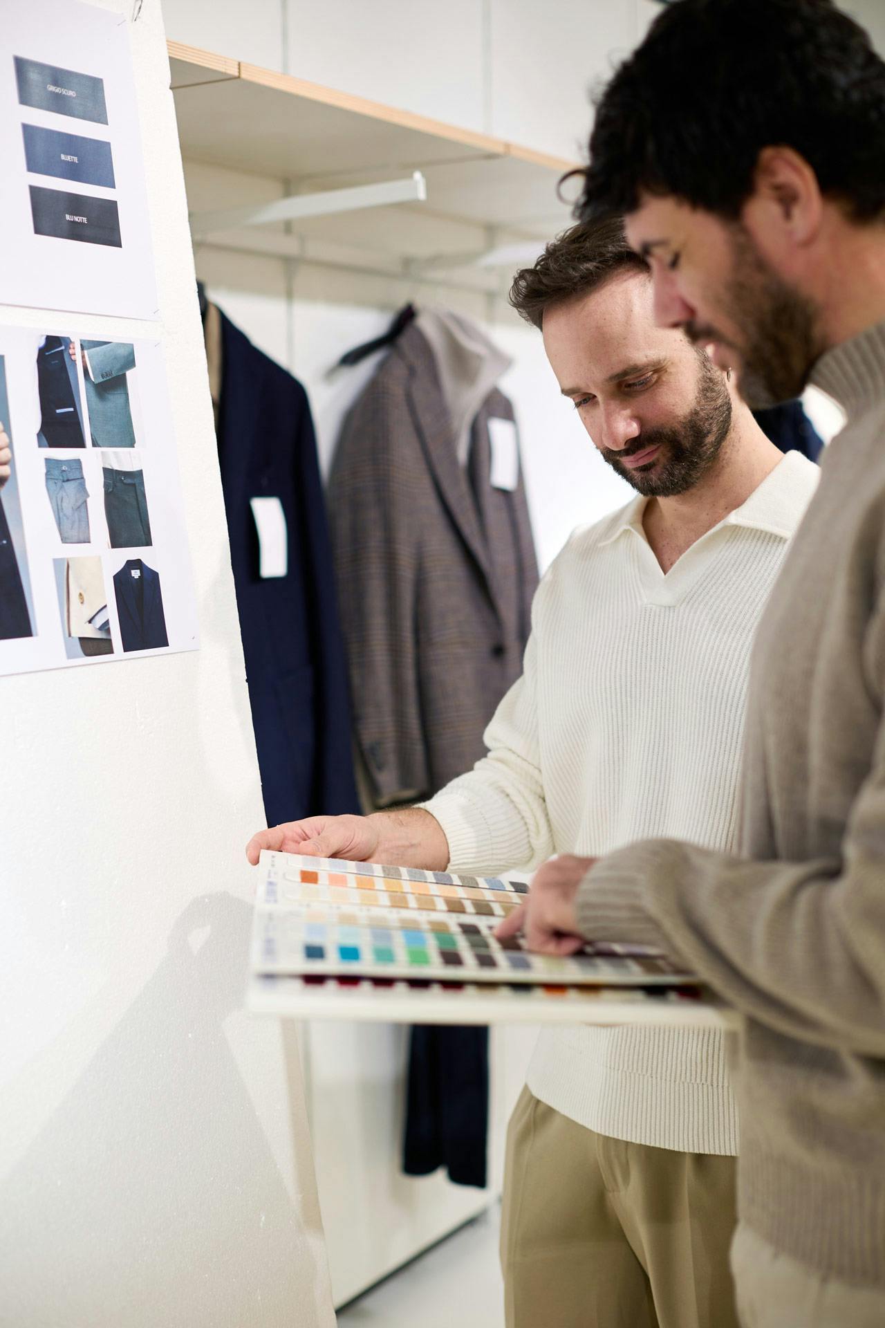 Due ragazzi esaminano palette colori in showroom moda maschile, campioni tessuto e abiti scuri appesi in background