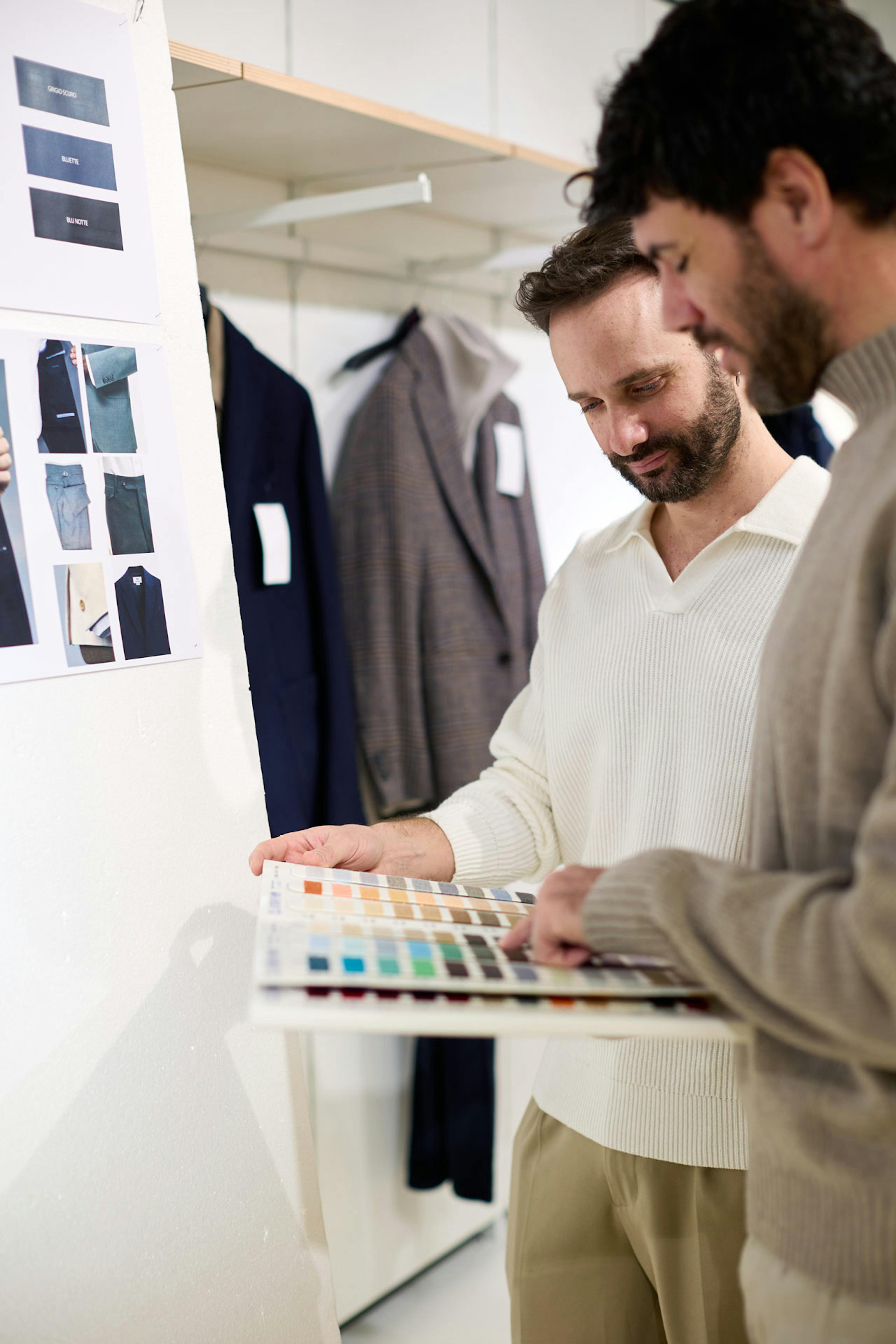 Due ragazzi esaminano palette colori in showroom moda maschile, campioni tessuto e abiti scuri appesi in background