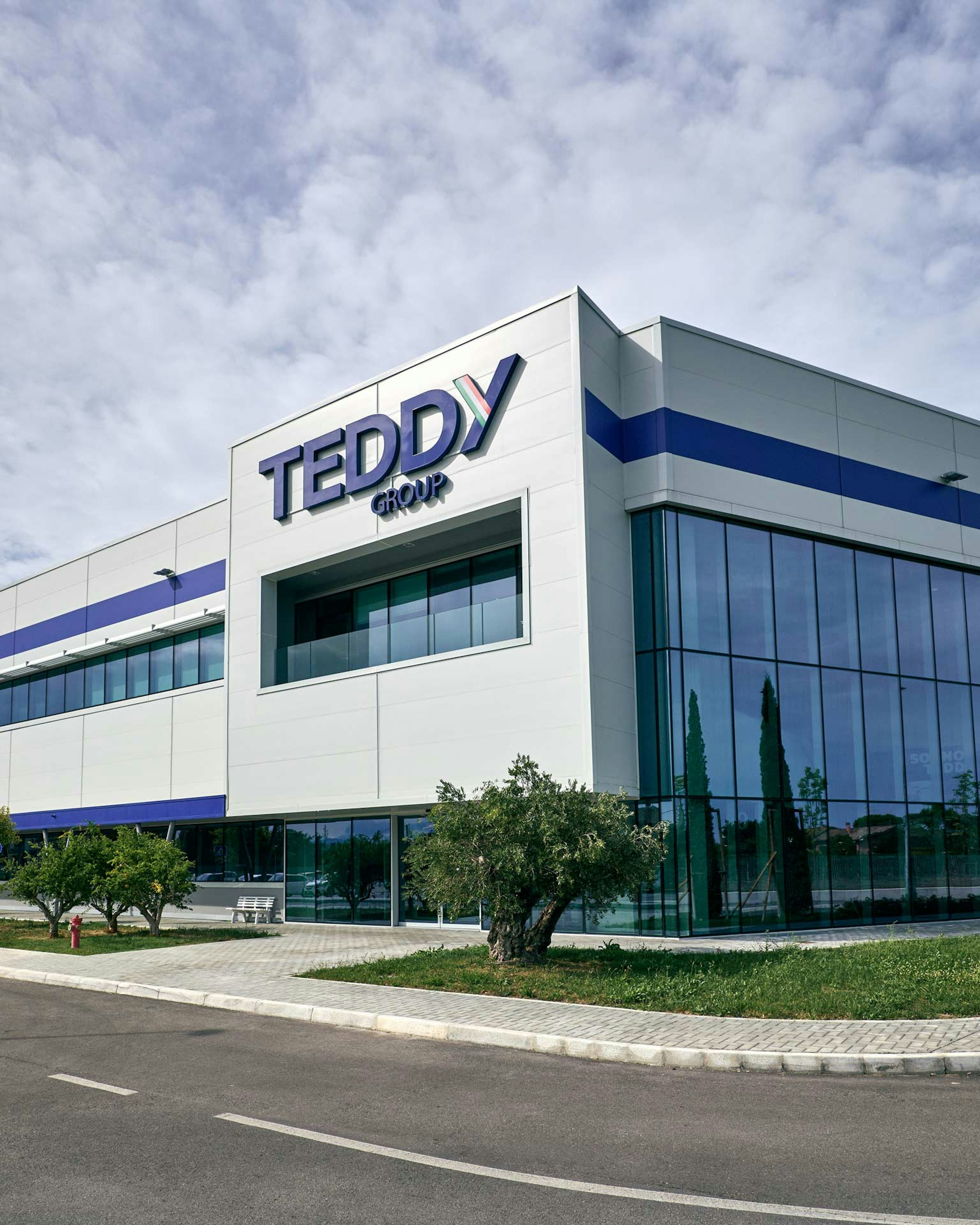 Edificio sede Teddy Group con architettura moderna, facciata bianca e dettagli blu, ampie vetrate e logo aziendale, cielo nuvoloso e area verde circostante