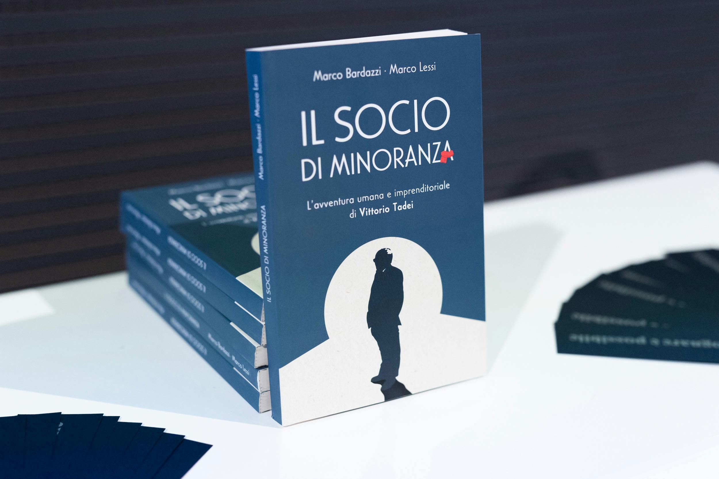 Pile di copie del libro 'Il socio di minoranza' su Vittorio Tadei disposte su tavolo bianco, copertina blu con figura stilizzata