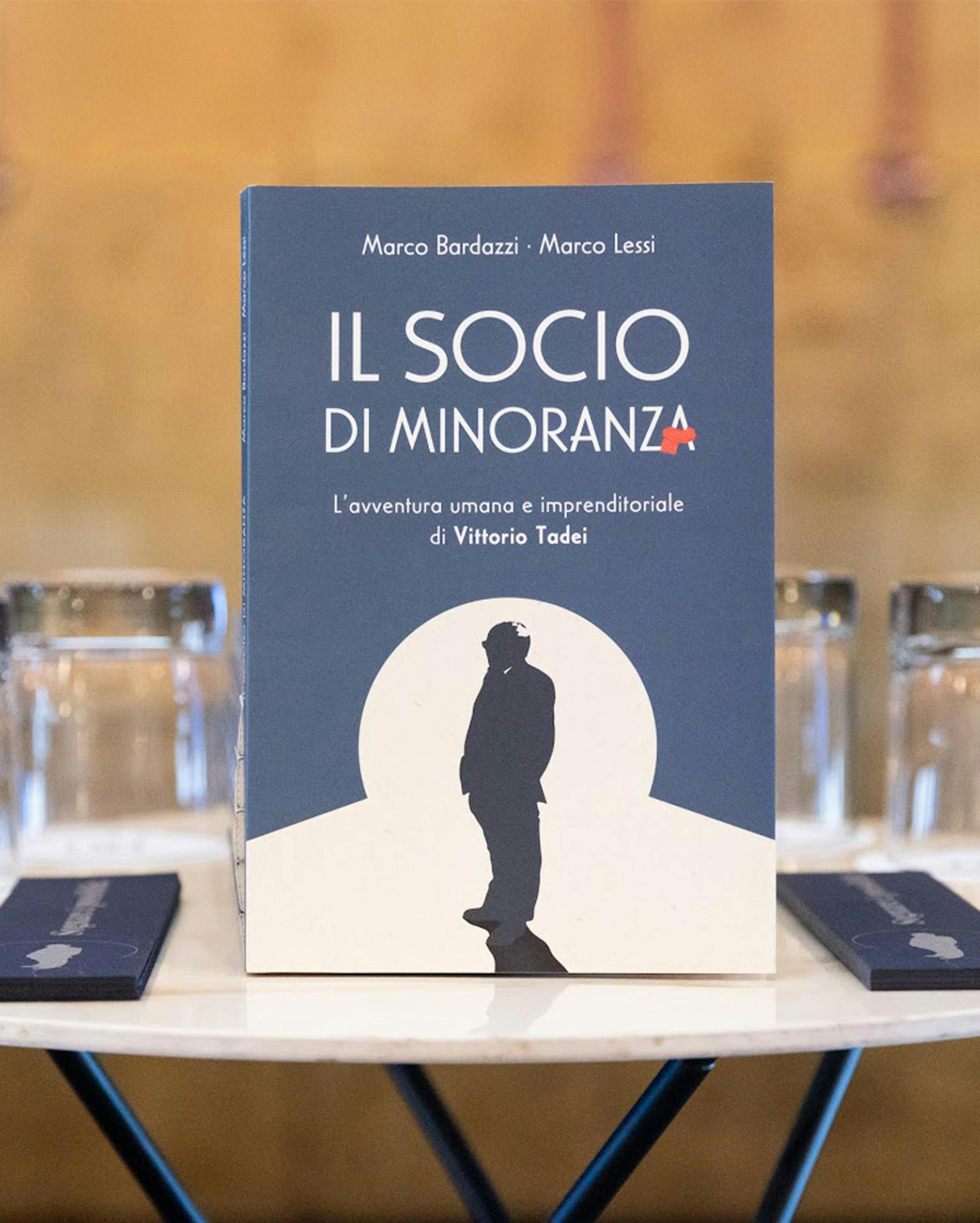 Libro 'Il socio di minoranza' di Marco Bardazzi e Marco Lessi su Vittorio Tadei, copertina blu con silhouette, esposto su tavolo con bicchieri