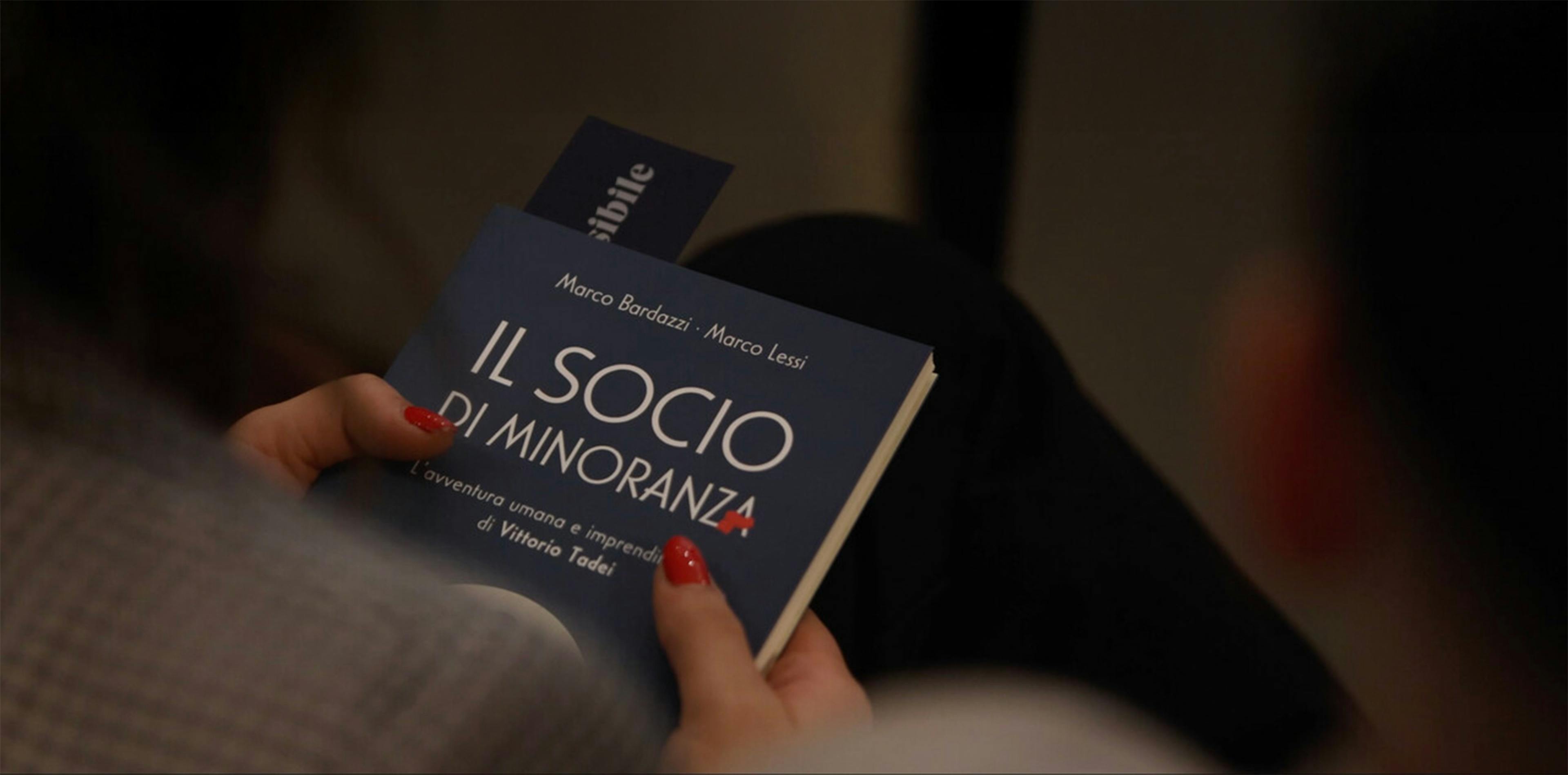 Mani femminili con smalto rosso tengono aperto il libro 'Il socio di minoranza' di Bardazzi e Lessi su Vittorio Tadei, segnalibro blu visibile