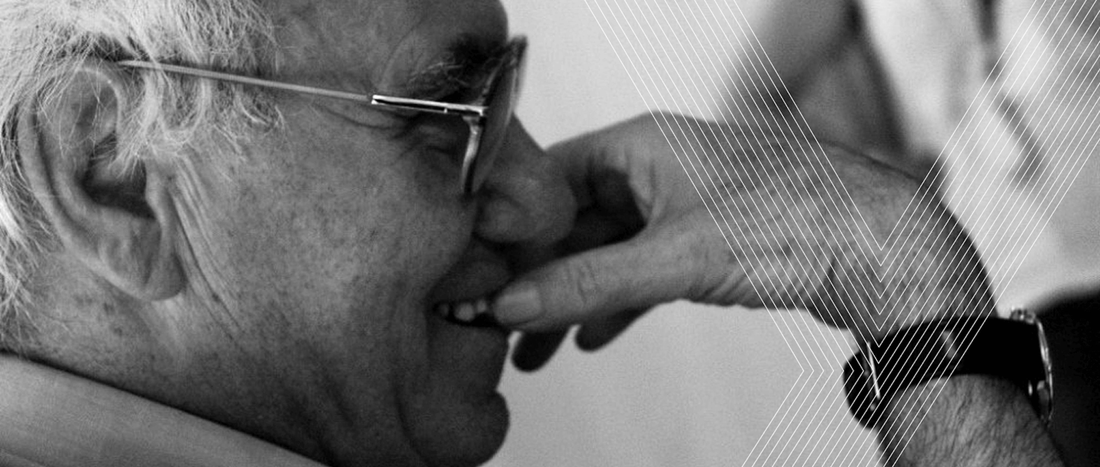 Ritratto in bianco e nero di Vittorio Tadei con occhiali che si porta la mano al viso in un gesto contemplativo. L'immagine presenta elementi grafici lineari sovrapposti che creano un effetto visivo dinamico