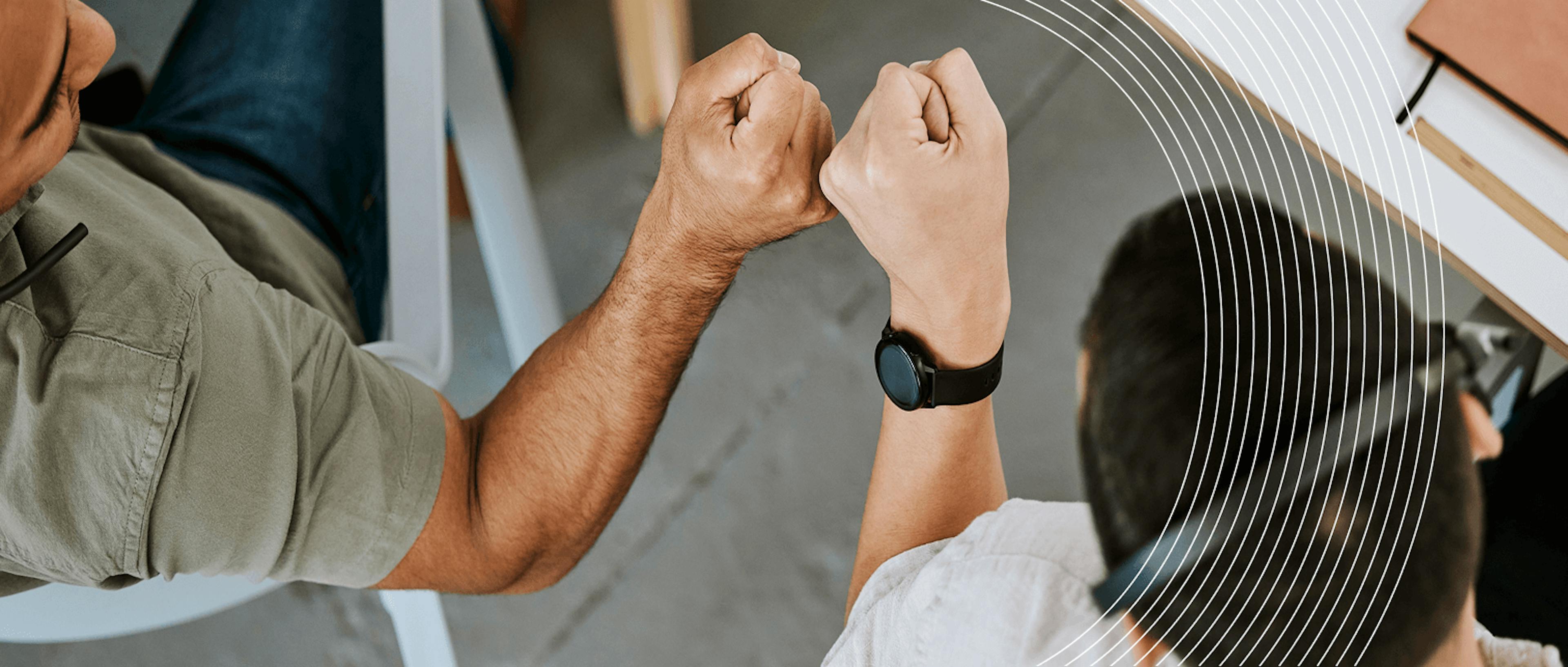 Due persone si tengono per mano in un gesto di connessione e supporto. L'immagine mostra solo le mani intrecciate, con uno dei partecipanti che indossa un orologio nero. La composizione include elementi grafici lineari che attraversano l'immagine