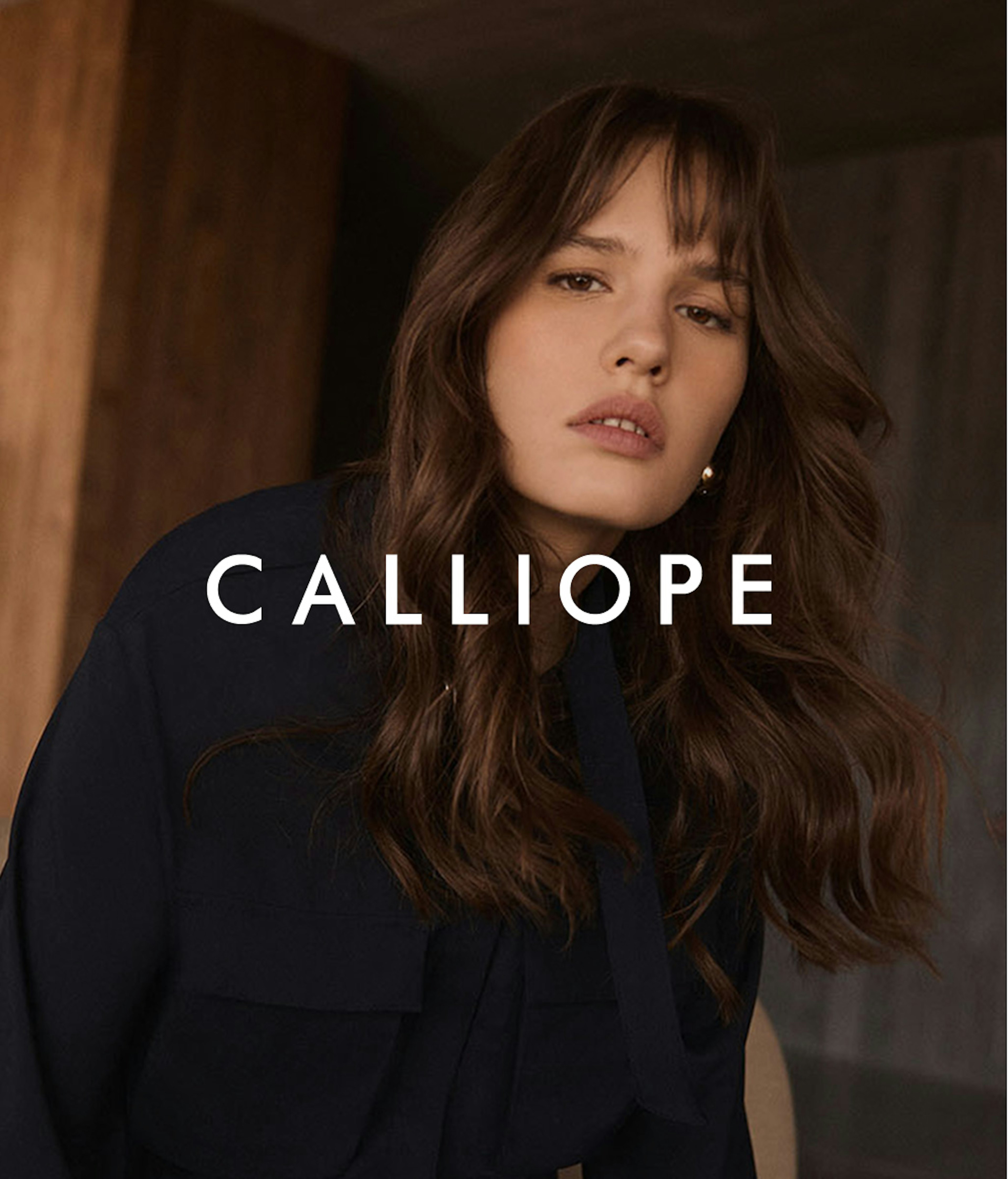 Modella con lunghi capelli castani mossi che indossa un blazer nero, fotografata in un ambiente interno con tonalità calde. Il nome del brand Calliope appare in caratteri bianchi al centro dell'immagine.