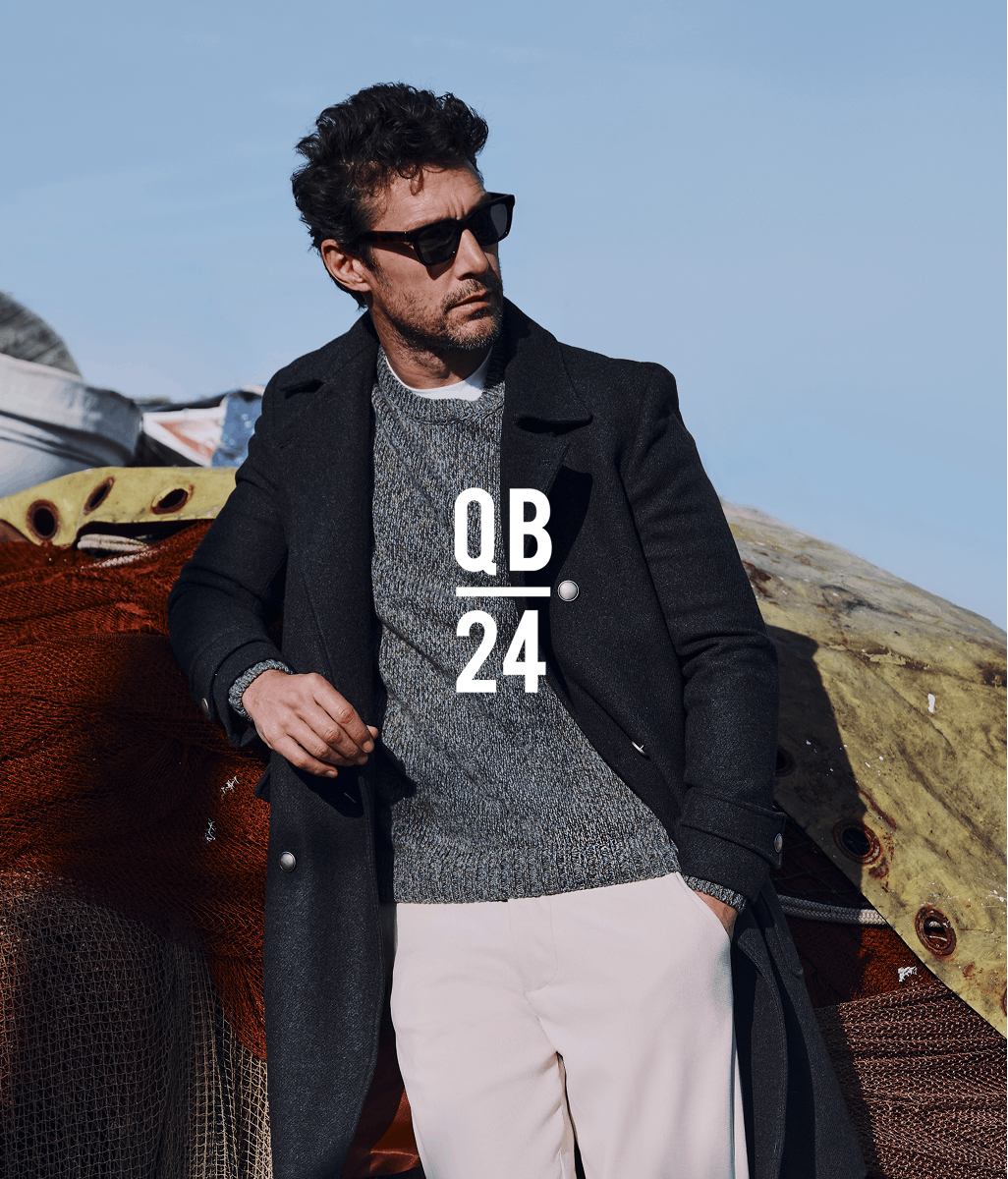 Modello in occhiali da sole, cappotto nero di lana sopra maglione grigio e pantaloni chiari, appoggiato a tessuto texturizzato all'aperto con montagne sullo sfondo. Testo sovrapposto 'QB 24' in bianco