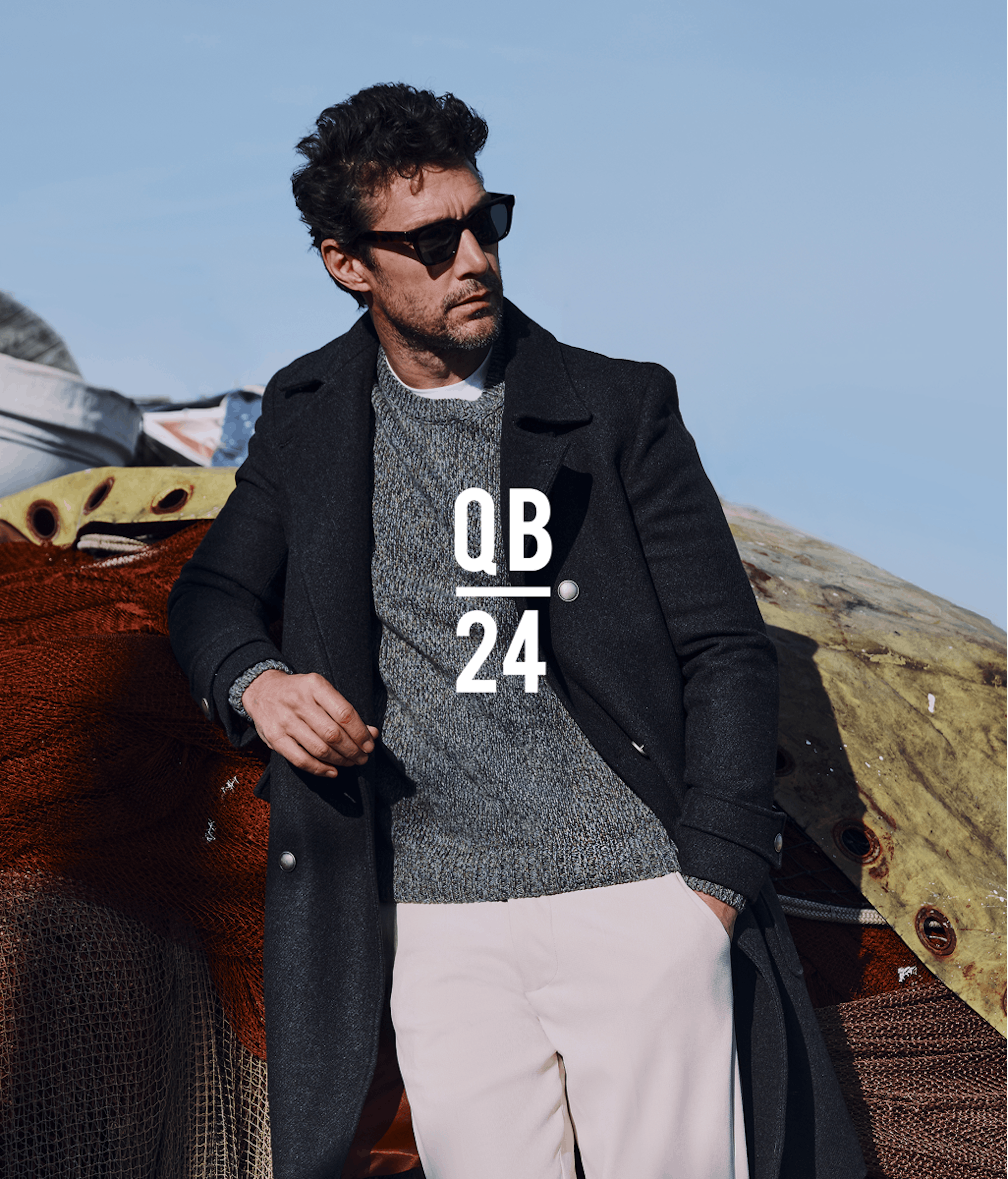 Modello in occhiali da sole, cappotto nero di lana sopra maglione grigio e pantaloni chiari, appoggiato a tessuto texturizzato all'aperto con montagne sullo sfondo. Testo sovrapposto 'QB 24' in bianco