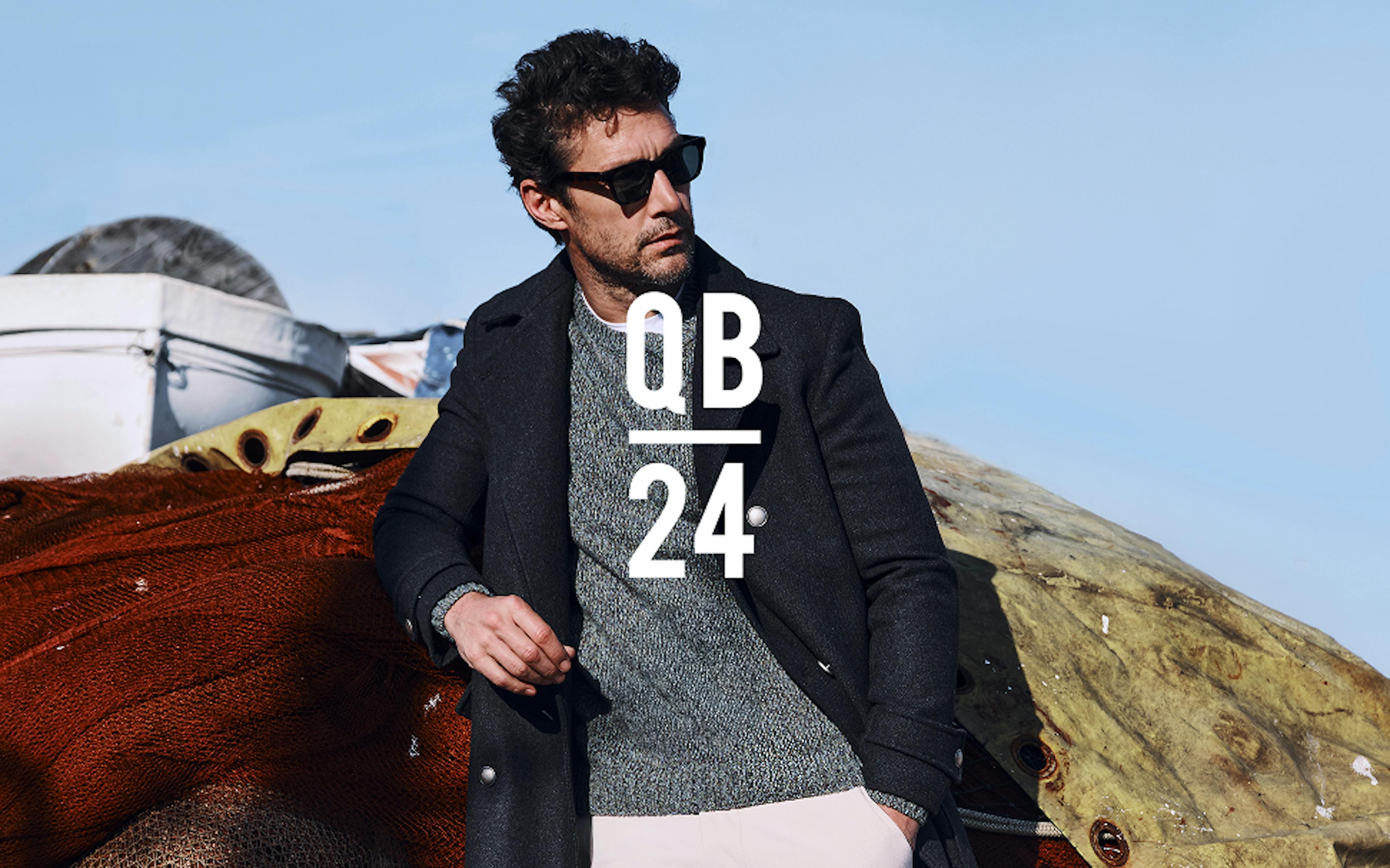 Modello in occhiali da sole, cappotto nero di lana sopra maglione grigio e pantaloni chiari, appoggiato a tessuto texturizzato all'aperto con montagne sullo sfondo. Testo sovrapposto 'QB 24' in bianco