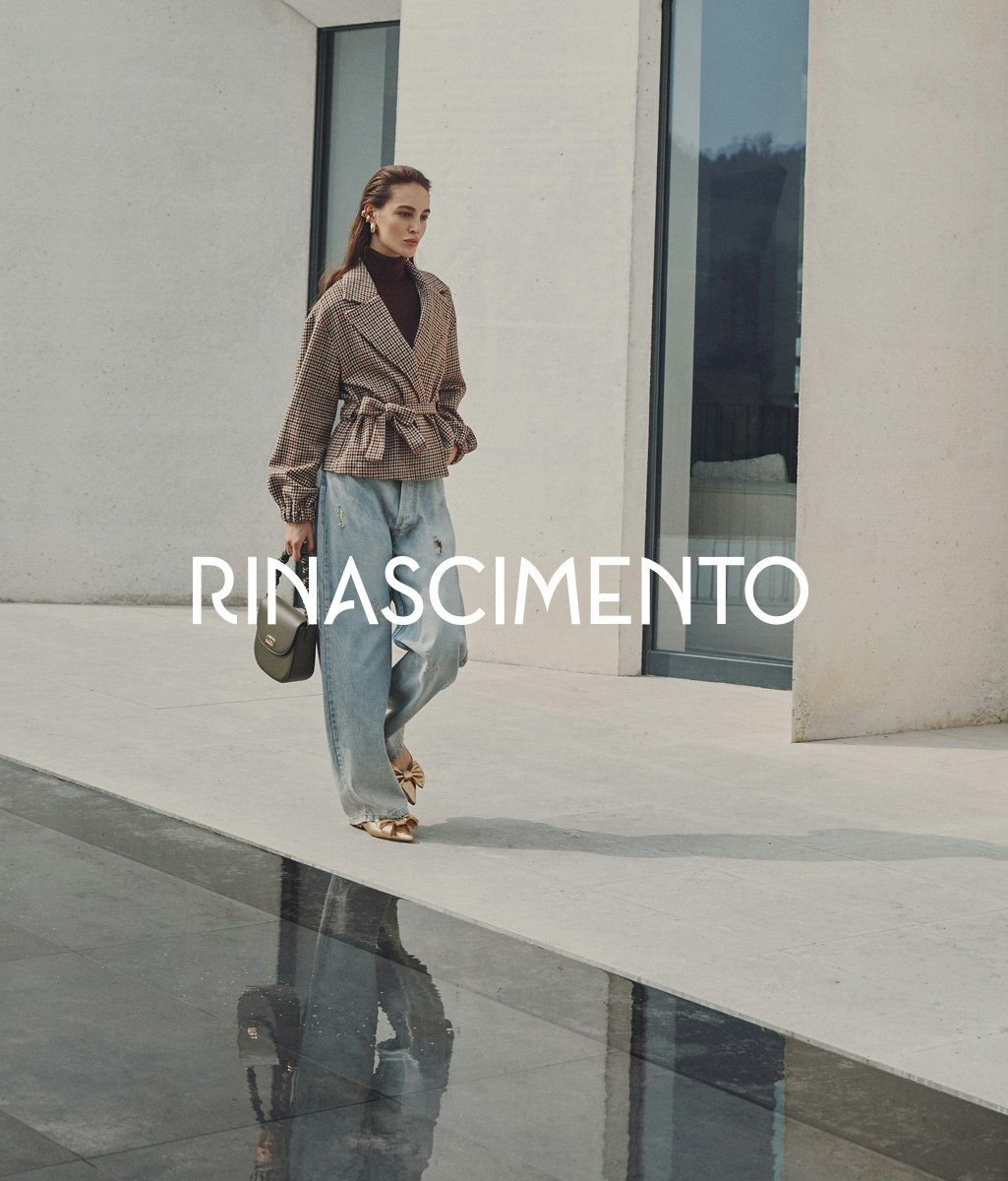 Modella in elegante completo beige composto da top senza maniche e pantaloni a gamba larga con cintura, che tiene una borsa beige, in posa contro elementi architettonici in pietra. Il nome Rinascimento appare in lettere bianche