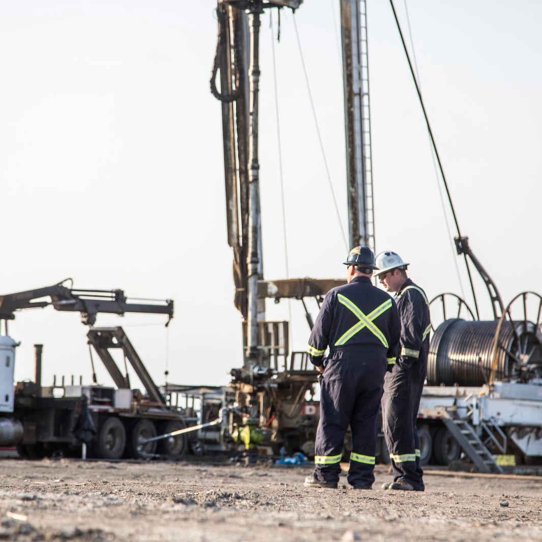 Two men in sunglasses and safety gear, speaking in front of an oil rig.